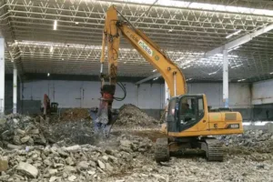 Escavadeira com rompedor demolindo estruturas dentro de galpão, com entulho separado no piso