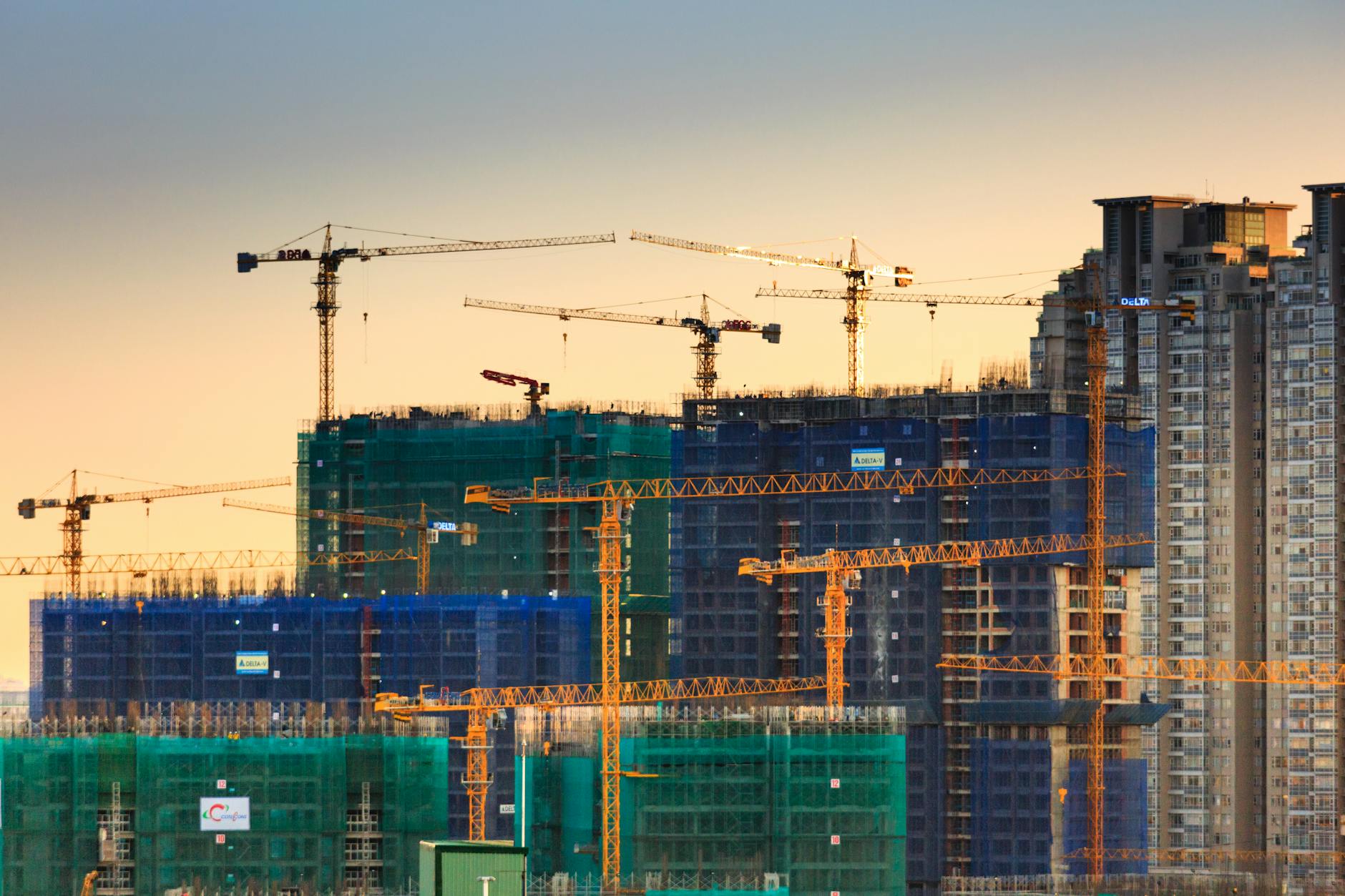 Prédios em construção com telas de proteção e várias gruas em obra de grande porte ao entardecer
