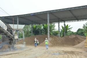 Engenheiros com capacete e colete caminham em pátio de obra com pilhas de areia sob cobertura metálica