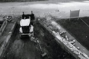 Retroescavadeira em escavação de terreno ao lado de muro de concreto demolido, em foto preto e branco