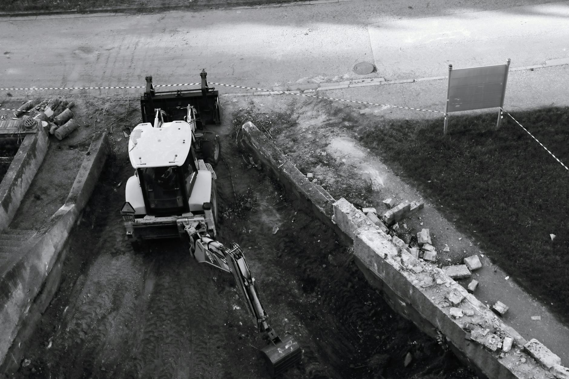 Retroescavadeira em escavação de terreno ao lado de muro de concreto demolido, em foto preto e branco
