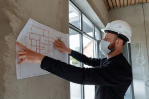 Engenheiro com capacete analisa planta de reforma de apartamento fixada na parede da obra