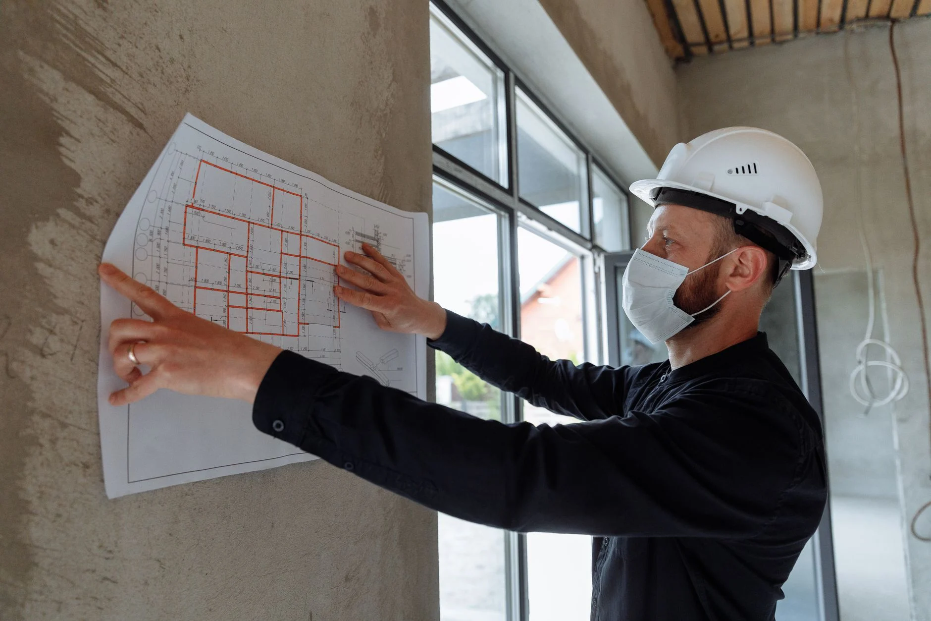 Engenheiro com capacete analisa planta de reforma de apartamento fixada na parede da obra
